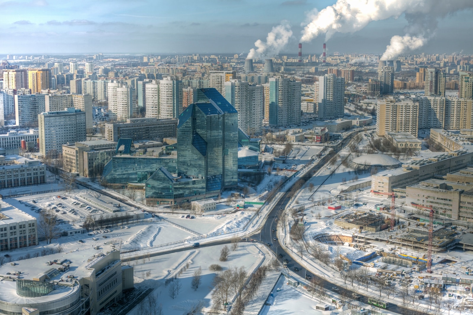 A winter view of Moscow’s Nikulino district from a 100-meter-tall rooftop. Fresh white snow blankets everything except the roads, with an abandoned building in the foreground and a thermal power plant about four kilometers away. It features two sets of cooling towers and two tall chimneys, all releasing thick steam into the cold sky.