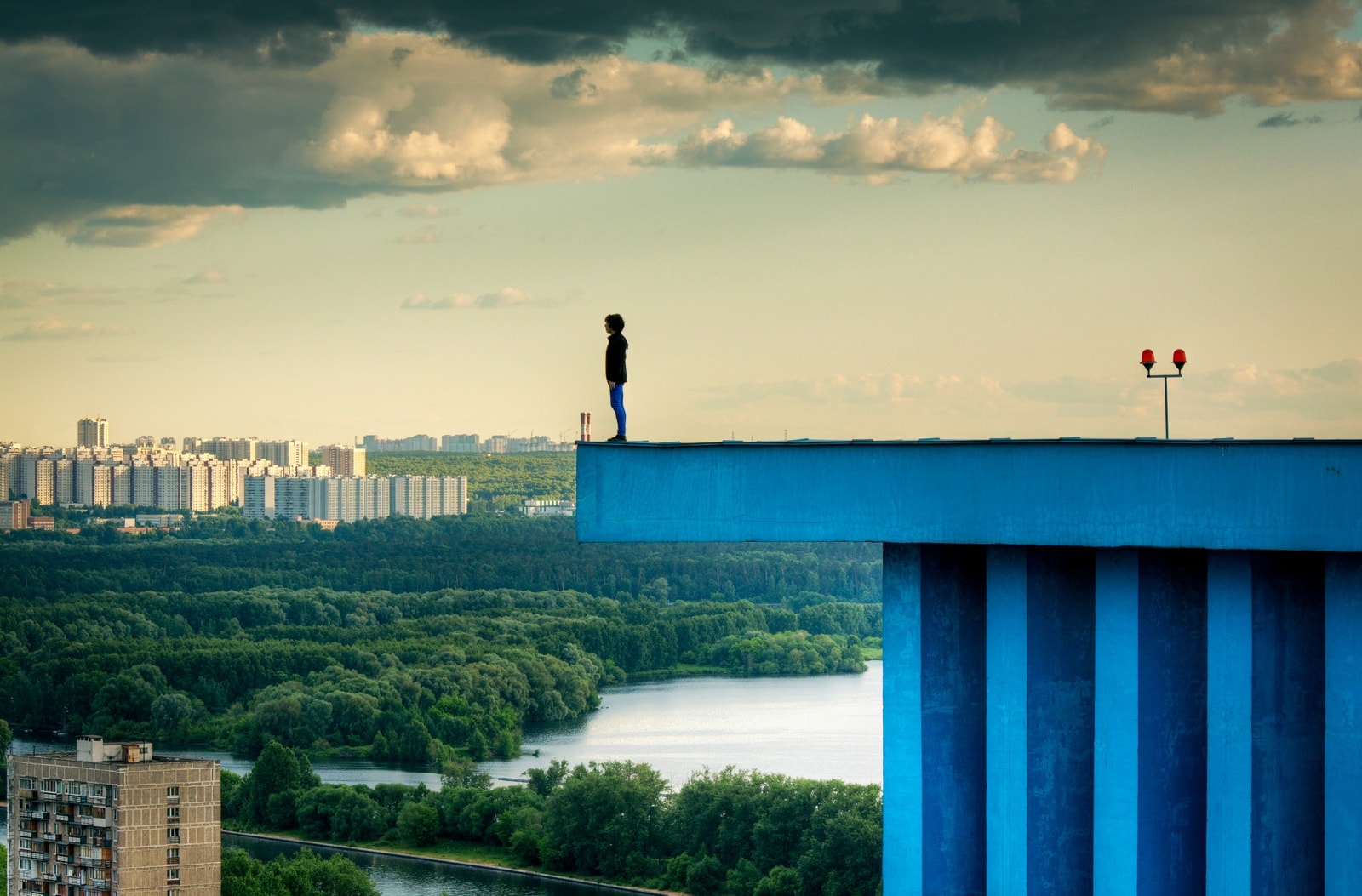 A young man stands motionless on the very edge of a tall rooftop in Moscow’s Shchukino district. Shot with ~130mm equivalent focal length; vibrant colors — saturated blue rooftop against a greenish sky.